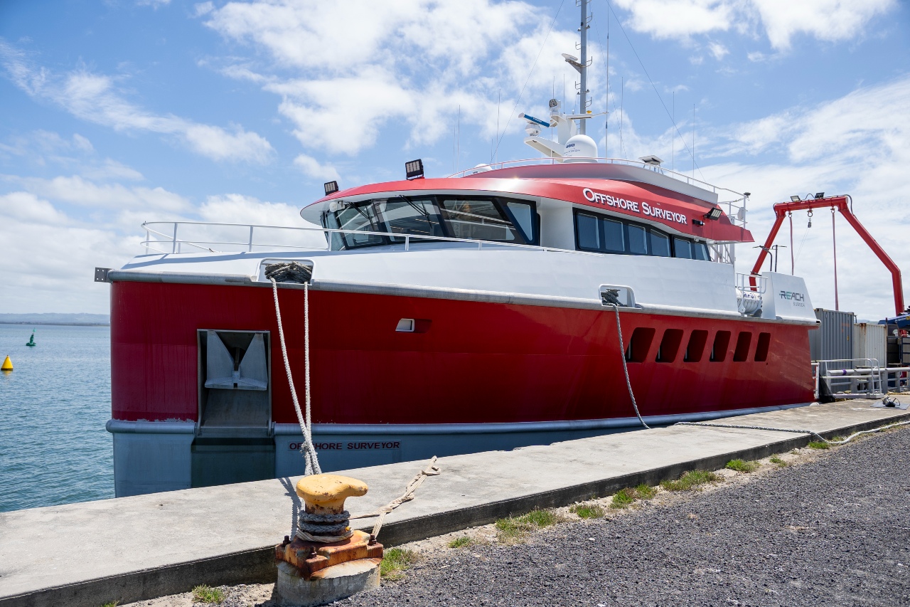 Red and white geophysical vessel named Offshore Surveyor docked at a pier under a partly cloudy sky