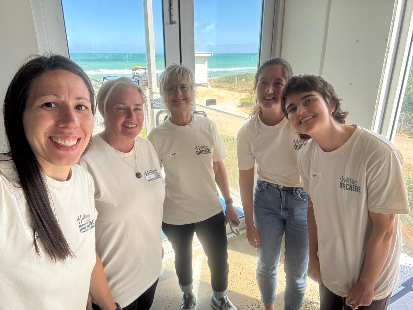 Five women smiling indoors with ocean view behind them, wearing Blue Mackerel shirts