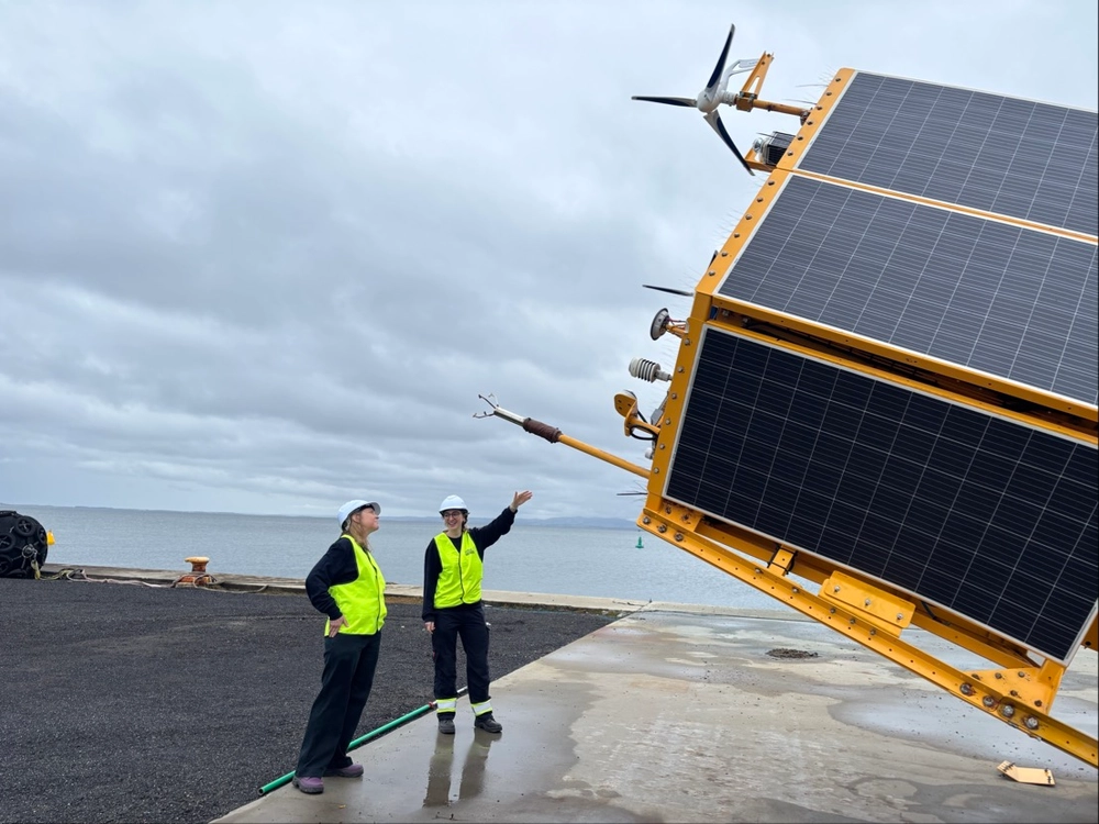 Two workers in high-visibility vests and helmets standing near a large solar panel installation by the water